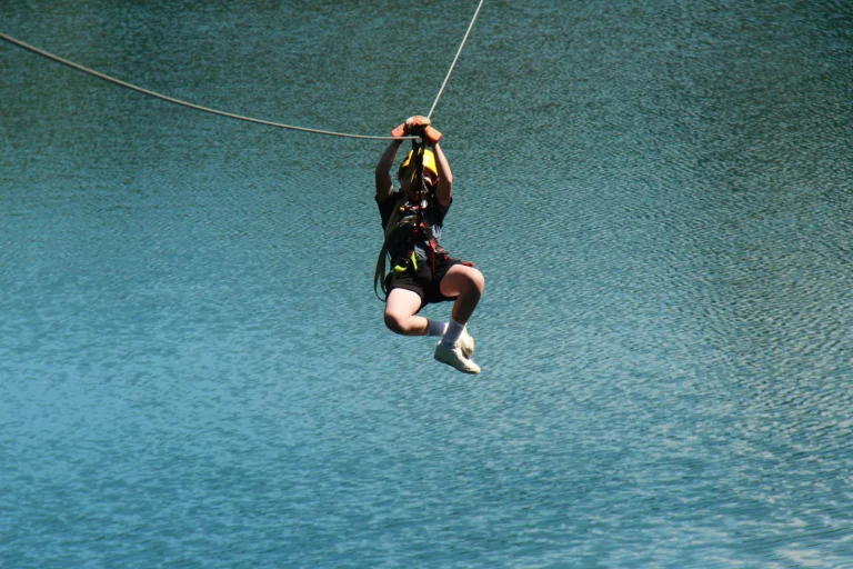 A camper riding a zip line high above clear blue-green water