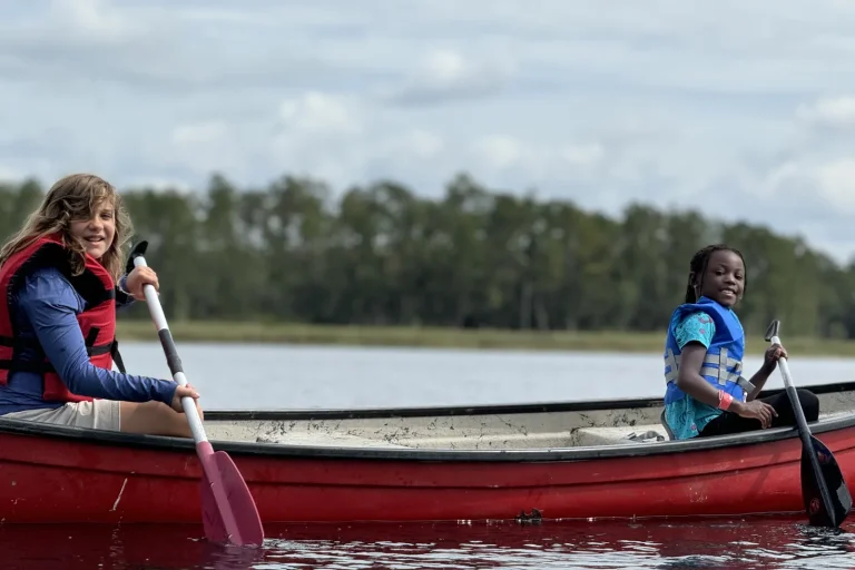 Two young campers in life jackets paddling a red canoe on the lake at Luther Springs