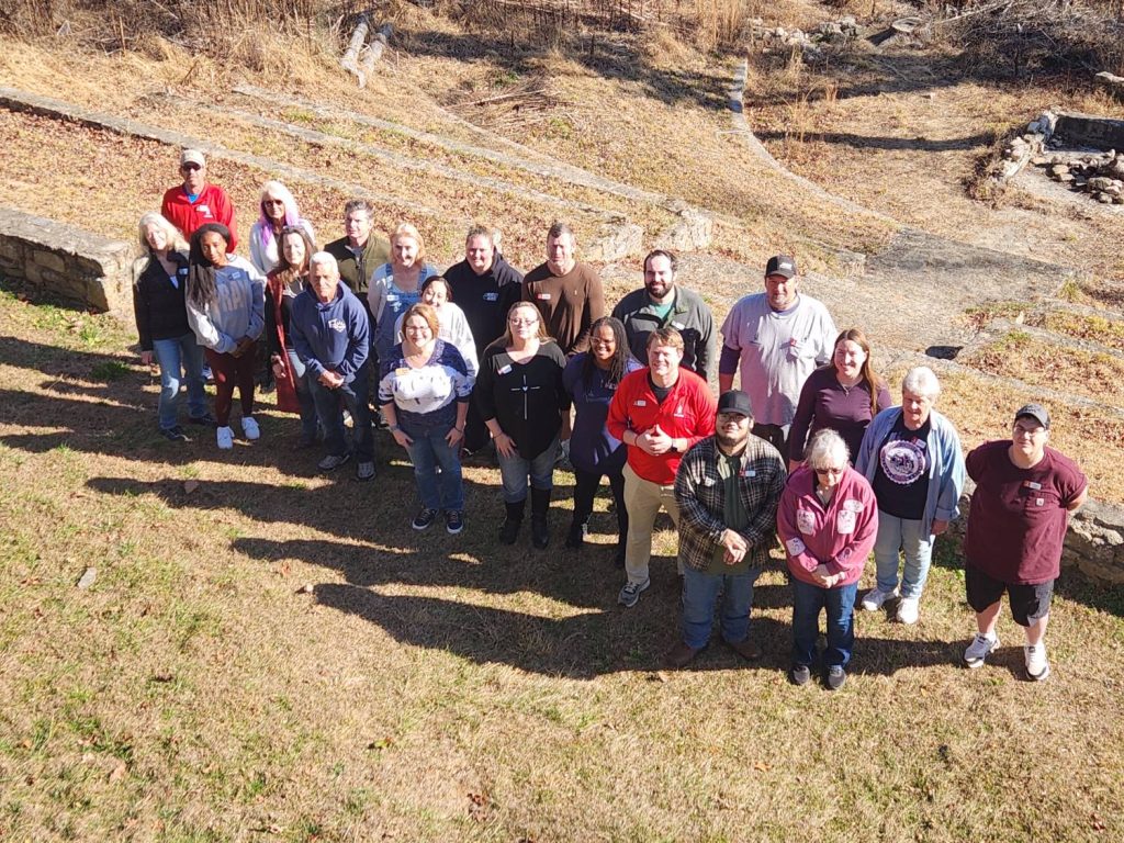 The NovusWay staff together at a staff retreat, gathered on a sunny hillside below an outdoor stone amphitheater