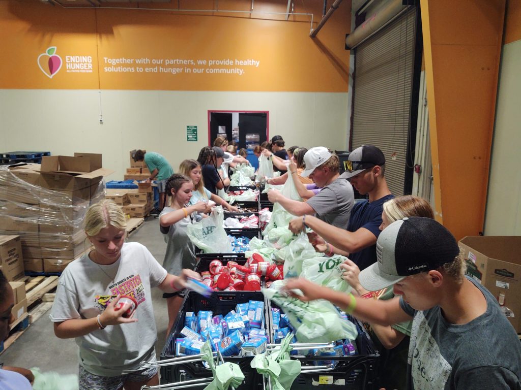 Volunteers pack bags of food at long tables in a food bank warehouse
