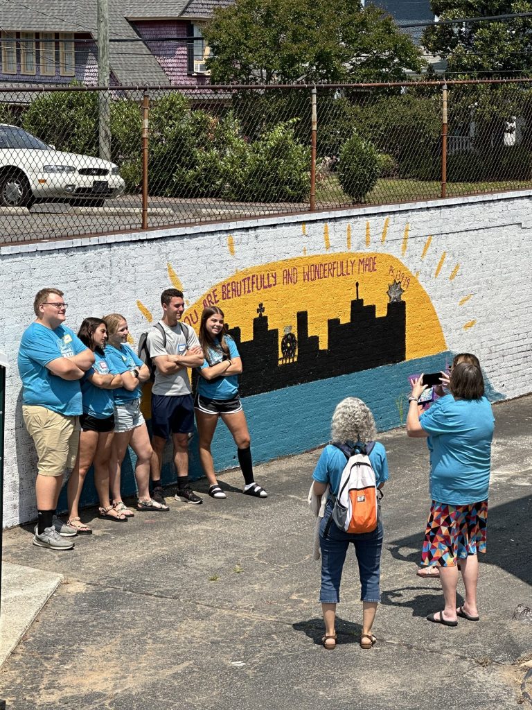 Young adults in matching blue T-shirts pose by a city mural that reads “You are beautifully and wonderfully made” while a woman takes their photo