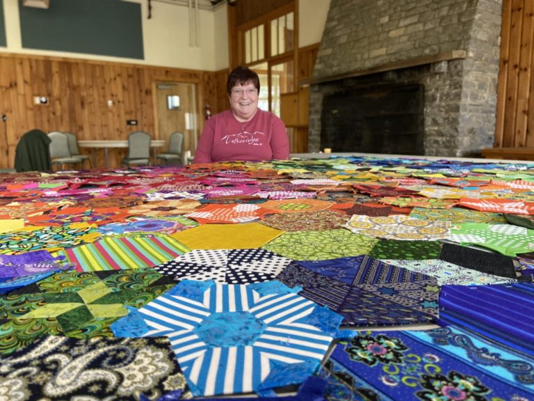 A smiling quilter behind a table covered in colorful hexagon quilt blocks