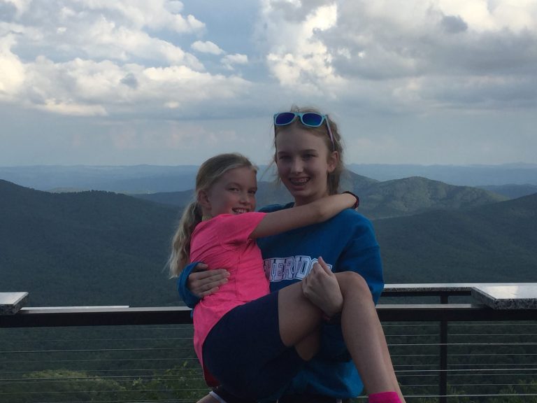 A smiling girl in a Lutheridge sweatshirt carries a younger girl at a mountain overlook
