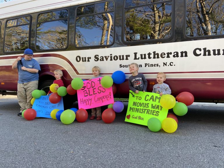 Young children with balloons and hand-lettered signs stand beside the Our Saviour Lutheran Church bus as it arrives at Lutheridge