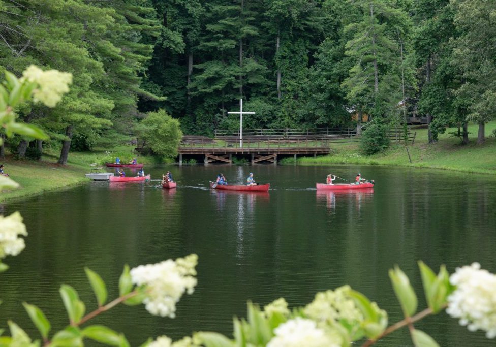 Campers paddle red canoes across the lake at Lutheridge, below the white cross on the hillside